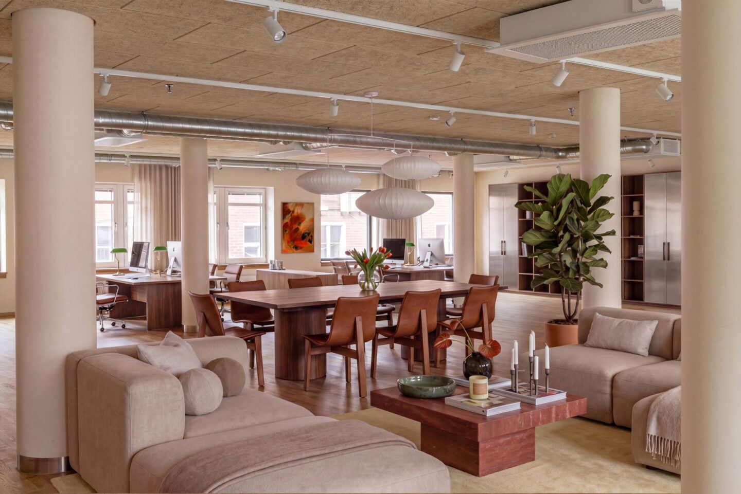 Modernt och stilrent kontor med skandinavisk inredning, öppna arbetsytor och loungegrupp i beige och trä. Lokalen har stora fönster, ljusa material, växter och designlampor över ett stort konferensbord, medan synliga ventilationsrör i taket ger en modern industriell känsla.