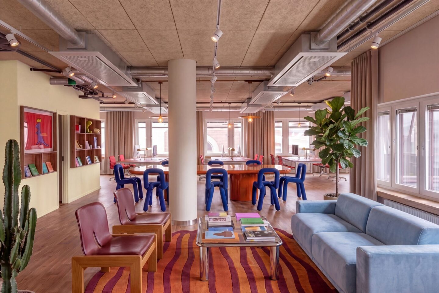Färgstarkt och modernt kontor med öppna arbetsytor, blå designstolar, soffgrupp och stora fönster som släpper in dagsljus. Lokalen har synliga ventilationsrör i taket, trädetaljer, växter och en kreativ inredning med böljande matta och konst på väggarna.