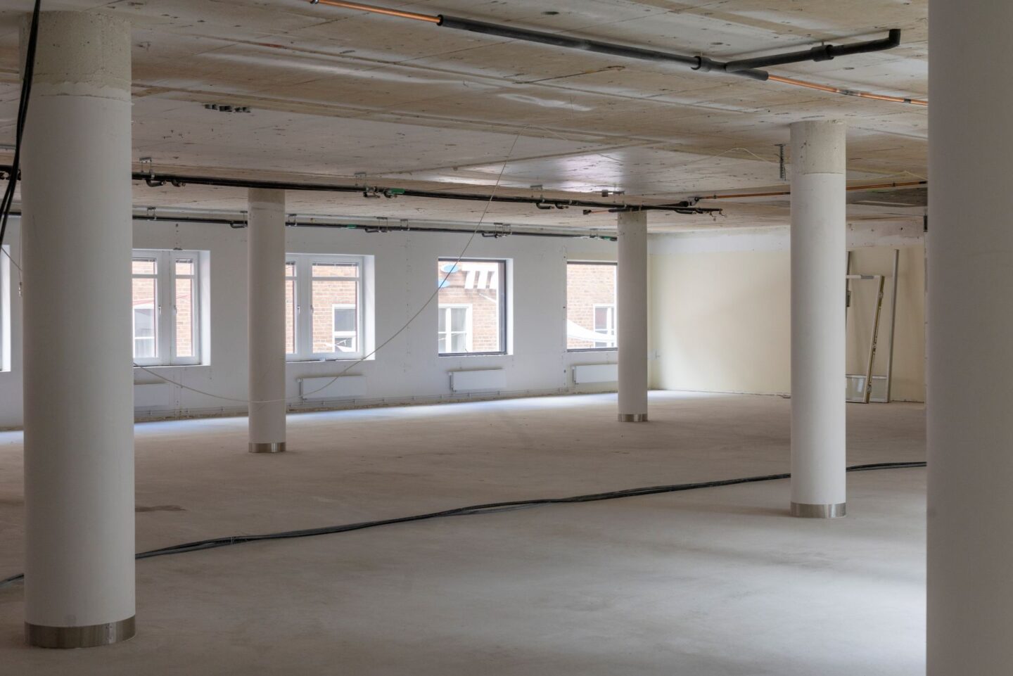 Stor öppen lokal under byggnation med vita pelare, synliga rör och kablar i taket samt stora fönster som släpper in dagsljus. Det tomma rummet har ljusa golv och en industriell känsla med pågående installationsarbete.