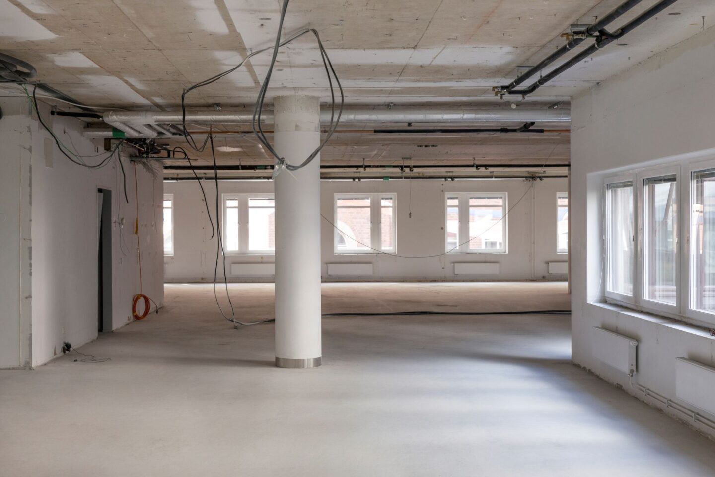 Stor öppen lokal under byggnation med betongtak, pelare och synliga installationer i taket. Lösa kablar hänger ner från taket och stora fönster längs väggarna släpper in dagsljus över det ljusa golvet och det tomma rummet.