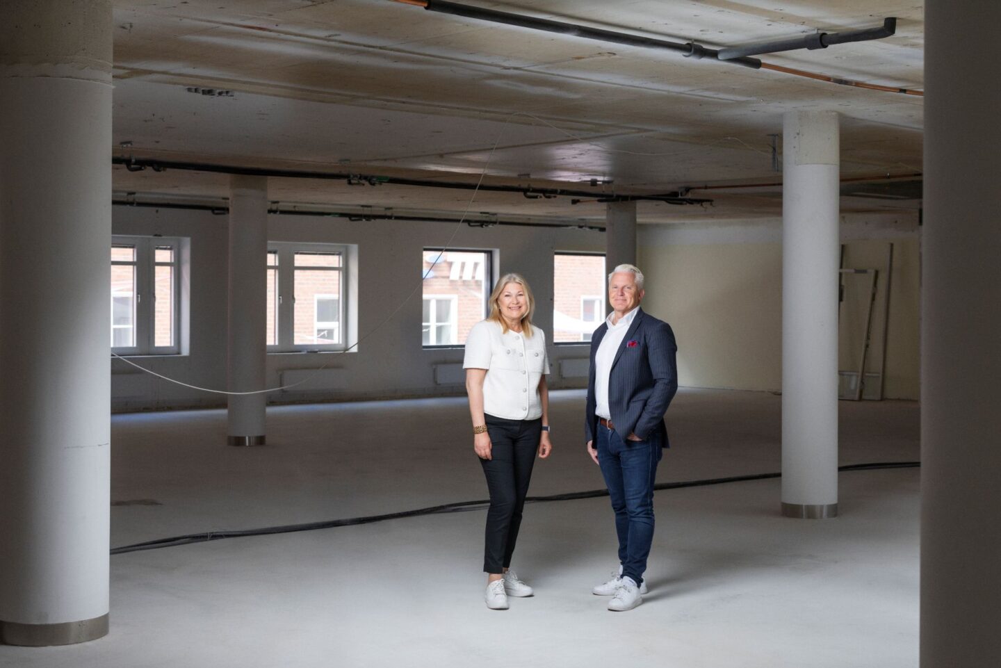 Två personer står i en stor, ljus lokal under byggnation med synliga pelare, takrör och stora fönster i bakgrunden. Personerna står mitt i rummet och bär vardagligt affärsklädda kläder. Det öppna utrymmet har ljusa golv och industriell känsla.