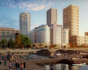 Alt-text (svenska): Modernt stadsområde vid vattnet med flera höga bostads- och kontorsbyggnader i varierande höjd och arkitektur. I förgrunden syns trädäck, kajer och vattenbassänger där människor promenerar, sitter och deltar i aktiviteter. En liten båt färdas på vattnet till höger. Träd och planteringar ger området en grön prägel, och kvällssolen skapar ett varmt ljus över byggnaderna och den offentliga miljön.